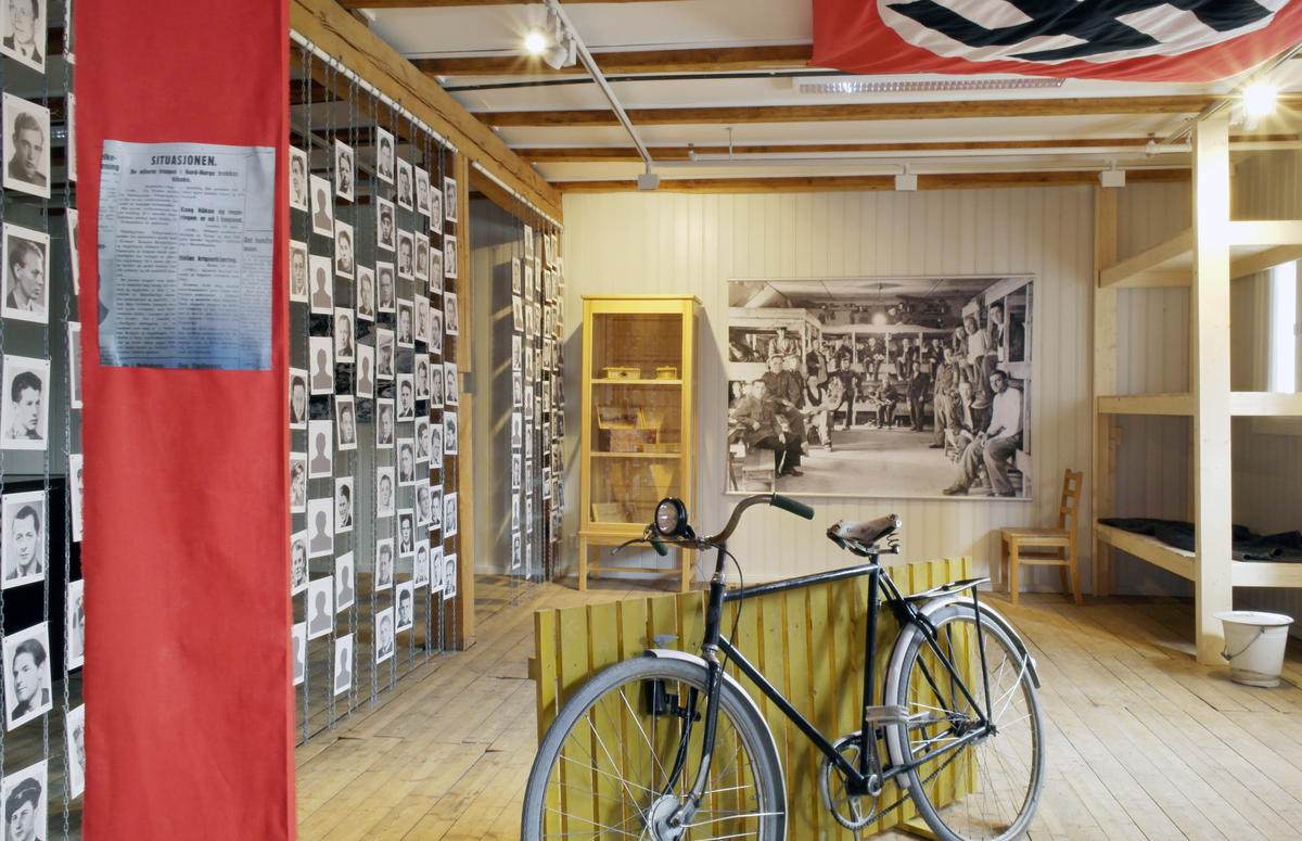 Bilde av utstillingen "Skudd i mørket - hva skjedde i Trandumskogen?" Rødt bånd med artikkel til venstre, sykkel på midten, brakkeseng til høyre, bilder av henrettede menn helt til venstre, stort bilde av krigsfanger og et monter med gjenstander bakers i rommet.