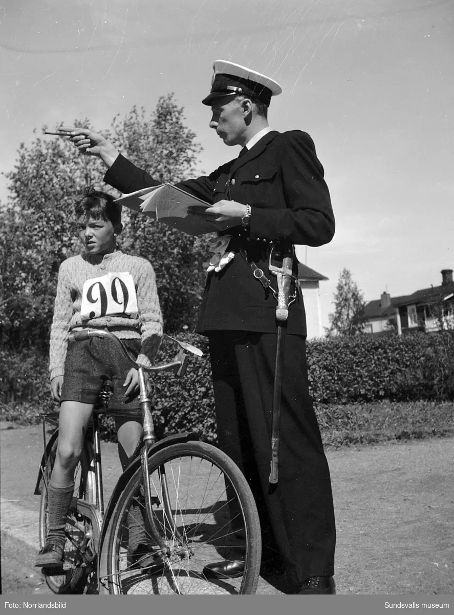 En poliskonstapel i uniform och en pojke på en cykel, pojken har nummerlapp på bröstet. Okänd adress.