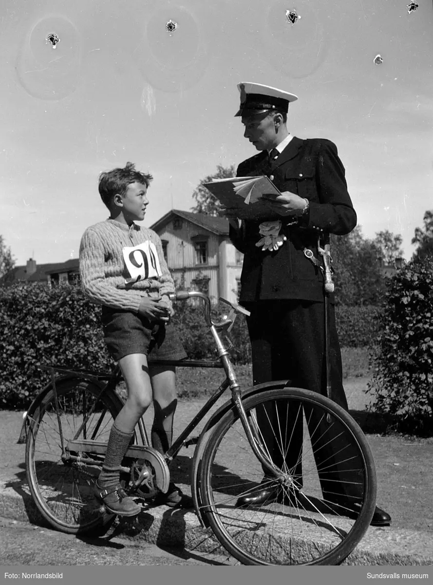 En poliskonstapel i uniform och en pojke på en cykel, pojken har nummerlapp på bröstet. Okänd adress.