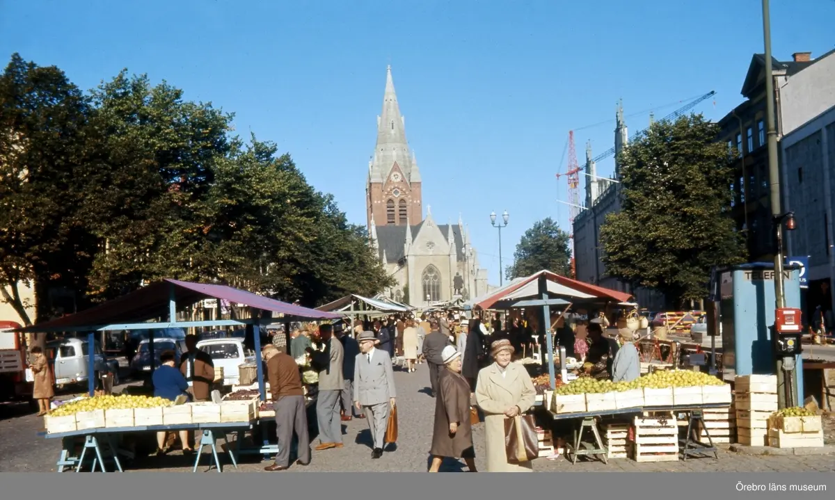 Stortorget med torghandel. Nikolaikyrkan och Engelbrektsstatyn i bakgrunden. 
Information från Trädgårdsmästare Börje Eriksson: torgståndet till vänster i bild är Arne Cederströms. Det är fru Cederström i blått. Torgståndet till höger är Rolf Cederströms, broder till Arne. Rolf står i ljusblå arbetsrock. 
Bilden är tagen före "Gamla normer, nya former"/"Likkistan", finns på Stortorget. 1960-tal.