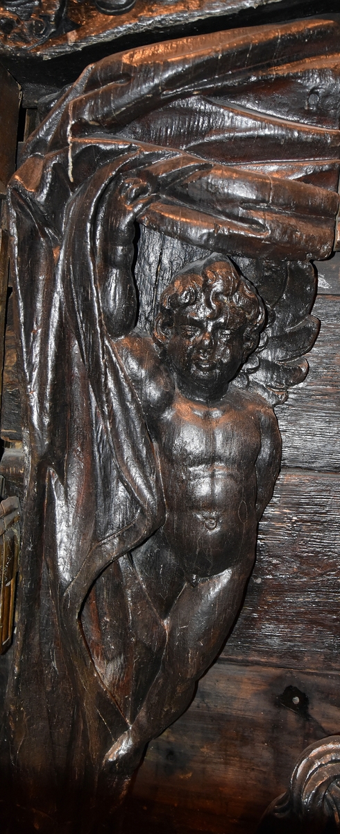 Putto samt större delen av ett draperi. Draperiet är kilformat med rak ytterkontur, och hålls undan av den svävande putton. Skulpturens baksida är slät.

Text in English: Putto and the larger part of a drapery. The drapery is wedge-shaped with straight outer contour, and is held back by the hovering putto. The reverse side is smooth.