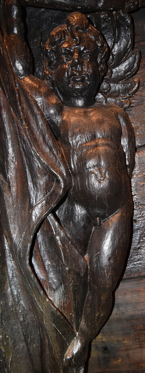 Putto samt större delen av ett draperi. Draperiet är kilformat med rak ytterkontur, och hålls undan av den svävande putton. Skulpturens baksida är slät.

Text in English: Putto and the larger part of a drapery. The drapery is wedge-shaped with straight outer contour, and is held back by the hovering putto. The reverse side is smooth.