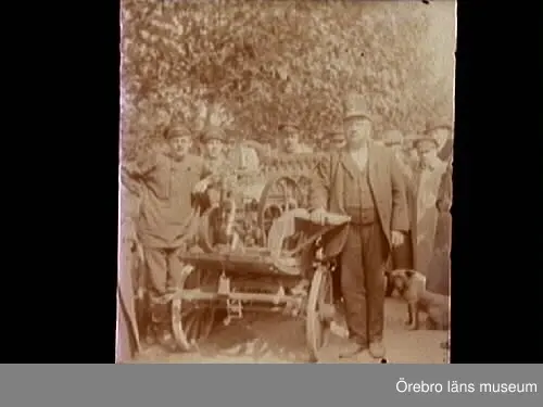 Män runt en bil. 
Fordonet på bilden är en hemmabyggd "automobil". Den är byggd i Gate, söder om Hjo, av smedmästaren Gustav Lindman och indelte soldaten Oscar Berg. Den är gjord av en gammal hästvagn och en encylindrig fotogenmotor, och sattes samman under natten till Kristi himmelsfärdsdag 1914. Den byggdes mest på skoj för att delta i ett "rally", men särskilt långt gick den dock inte. Den tog sig ett par kilometer från Gate smedja till Gate Handel, men sedan var sagan all.
Bilden är tagen vid Gate smedja. Gustav Lindman syns längst fram till vänster. Bakom sätet står Oscar Berg och mannen längst fram till höger är David Johansson från Skrinhult.