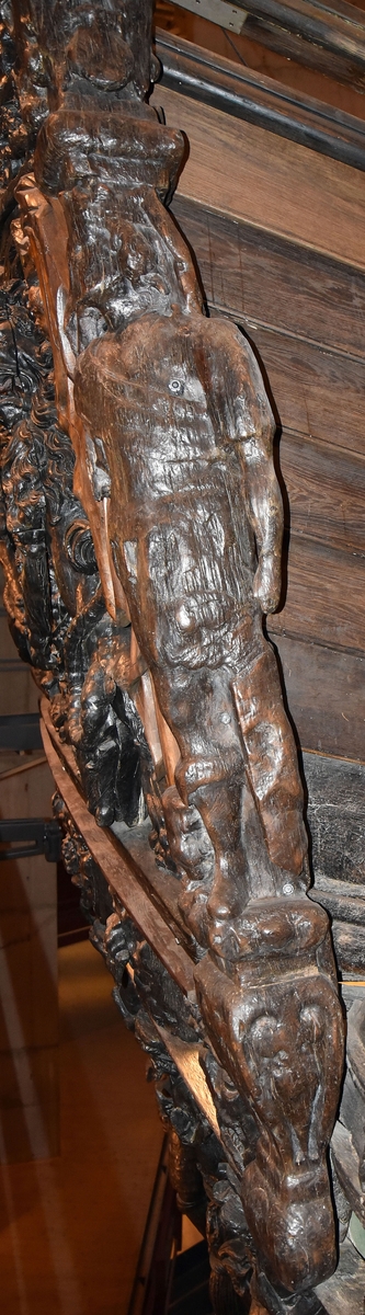 Skulptur i form av en hög, rikt snidad pelare eller stolpe, med kvadratisk genomskärning.

Tre av pelarens sidor är skulpterade medan den fjärde utgör anliggningsyta, försedd med ett antal olikformade urtag. De skulpterade sidorna är horisontellt indelade i tre eller fyra sektioner eller nivåer, avgränsade från varandra av profilerade och snedställda vulster. Nedtill avslutas skulpturen av en ornerad och på mitten insvängd formation.



Ovanför denna nedre avslutning är pelaren utformad som en reslig mansfigur, vänd mot vänster, sedd från betraktaren. Mansfiguren, som täcker tre av skulpturens fyra sidor, är iförd en romersk rustning med en bred fältbindel draperad från höger axel snett över bröstet. På huvudet med det lockiga och utslagna håret sitter en krona av öppen typ, med liljeliknande ornament. Figurens vänstra arm är böjd, medan den högra armen är sträckt utmed sidan. Handen till den högra armen saknas, men har hållit en rikt profilerad sköld, med mycket slitna och vaga konturer av en på bakbenen mot vänster gående grip med stora vingar. Skölden är snidad i ett med figuren, men med kraftiga underskärningar. Vid figurens fötter skymtar ett djur av något slag. Bakom mannens huvud tycks ett snidat textilstycke, kanske del till ett draperi, vara anbragt.



Ovanför den romerska krigaren eller riddaren har pelaren formen av en herm utan armar med långt, krusigt hår och mustaschprytt ansikte. Liksom mansfiguren täcker även hermen tre sidor av pelaren. Huvudet är vridet mot vänster. Överkroppen är bar så när som på ett flikigt plagg som täcker axlar och övre delen av bröstet. Från huvudet och runt midjan är ett tygstycke draperat. Hermens nederdel är formad till en stor maskaron med uppspärrad mun och utsträckt tunga. Maskaronen övergår upptill och utmed sidorna i yppiga bladformationer.



Pelaren avslutas upptill av ett knoppliknande ornament. Ornamentet är ornerat med akantusbladliknande bildningar samt fyra byglar som möts på toppen, där vissa spår antyder att det ursprungligen suttit ett klot eller dylikt, men som fallit bort. Eventuellt kan ornamentet utgöra en huvudbonad till hermen.

Skulpturen är kraftigt sliten.



Text in English: Sculpture in the shape of a richly carved column or pillar.

Three sides of the pillar are carved, while the fourth side comprises the attachment surface and has a number of variously-shaped indentations. The sculptured sides are divided horizontally into three or four sections separated by angled tori.



The lower section of the pillar is carved in the figure of a tall man. The figure is clad in Roman armour and has a field badge (cloak/plaid) draped across his chest at an angle from his right shoulder. On his head he wears an open crown. He has long, curly hair. The right arm is broken off, the left is at his side. The left hand is missing but once held a richly carved shield.

The shield is carved out of the same piece of wood as the figure, but deeply undercut. A carved figure is apparent on the shield. Although the shield is badly worn, it is possible to recognise one of the hind legs of a griffin with large wings. At the man's feet lies an animal of some kind.



Above the Roman warrior or knight, the pillar is carved in the shape of a herm, without arms and with long, curly hair and a moustache. Like the male figure, the herm also covers three sides of the pillar. The head is twisted to the left. The upper part of the body is bare except for a piece of cloth that covers the shoulders and upper chest. The lower part of the herm is carved as a large mask, its mouth open and tongue protruding. The sides and upper parts of the mask are carved as luxuriant leaves.



The upper part of the pillar has a ornament in shape of a knob. The ornament has a ring of acanthus leaves and four spherical loops run vertically from the acanthus leaves and meet at a point at the top. An object, for example a globe appears to have adorned the top of the ornament. This object is missing. The ornament may also be a part of the headgear of the herm.

The sculpture is severely worn.