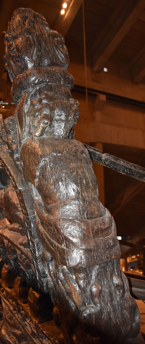 Skulptur i form av en hög, rikt snidad pelare eller stolpe, med kvadratisk genomskärning.

Tre av pelarens sidor är skulpterade medan den fjärde utgör anliggningsyta, försedd med ett antal olikformade urtag. De skulpterade sidorna är horisontellt indelade i tre eller fyra sektioner eller nivåer, avgränsade från varandra av profilerade och snedställda vulster. Nedtill avslutas skulpturen av en ornerad och på mitten insvängd formation.



Ovanför denna nedre avslutning är pelaren utformad som en reslig mansfigur, vänd mot vänster, sedd från betraktaren. Mansfiguren, som täcker tre av skulpturens fyra sidor, är iförd en romersk rustning med en bred fältbindel draperad från höger axel snett över bröstet. På huvudet med det lockiga och utslagna håret sitter en krona av öppen typ, med liljeliknande ornament. Figurens vänstra arm är böjd, medan den högra armen är sträckt utmed sidan. Handen till den högra armen saknas, men har hållit en rikt profilerad sköld, med mycket slitna och vaga konturer av en på bakbenen mot vänster gående grip med stora vingar. Skölden är snidad i ett med figuren, men med kraftiga underskärningar. Vid figurens fötter skymtar ett djur av något slag. Bakom mannens huvud tycks ett snidat textilstycke, kanske del till ett draperi, vara anbragt.



Ovanför den romerska krigaren eller riddaren har pelaren formen av en herm utan armar med långt, krusigt hår och mustaschprytt ansikte. Liksom mansfiguren täcker även hermen tre sidor av pelaren. Huvudet är vridet mot vänster. Överkroppen är bar så när som på ett flikigt plagg som täcker axlar och övre delen av bröstet. Från huvudet och runt midjan är ett tygstycke draperat. Hermens nederdel är formad till en stor maskaron med uppspärrad mun och utsträckt tunga. Maskaronen övergår upptill och utmed sidorna i yppiga bladformationer.



Pelaren avslutas upptill av ett knoppliknande ornament. Ornamentet är ornerat med akantusbladliknande bildningar samt fyra byglar som möts på toppen, där vissa spår antyder att det ursprungligen suttit ett klot eller dylikt, men som fallit bort. Eventuellt kan ornamentet utgöra en huvudbonad till hermen.

Skulpturen är kraftigt sliten.



Text in English: Sculpture in the shape of a richly carved column or pillar.

Three sides of the pillar are carved, while the fourth side comprises the attachment surface and has a number of variously-shaped indentations. The sculptured sides are divided horizontally into three or four sections separated by angled tori.



The lower section of the pillar is carved in the figure of a tall man. The figure is clad in Roman armour and has a field badge (cloak/plaid) draped across his chest at an angle from his right shoulder. On his head he wears an open crown. He has long, curly hair. The right arm is broken off, the left is at his side. The left hand is missing but once held a richly carved shield.

The shield is carved out of the same piece of wood as the figure, but deeply undercut. A carved figure is apparent on the shield. Although the shield is badly worn, it is possible to recognise one of the hind legs of a griffin with large wings. At the man's feet lies an animal of some kind.



Above the Roman warrior or knight, the pillar is carved in the shape of a herm, without arms and with long, curly hair and a moustache. Like the male figure, the herm also covers three sides of the pillar. The head is twisted to the left. The upper part of the body is bare except for a piece of cloth that covers the shoulders and upper chest. The lower part of the herm is carved as a large mask, its mouth open and tongue protruding. The sides and upper parts of the mask are carved as luxuriant leaves.



The upper part of the pillar has a ornament in shape of a knob. The ornament has a ring of acanthus leaves and four spherical loops run vertically from the acanthus leaves and meet at a point at the top. An object, for example a globe appears to have adorned the top of the ornament. This object is missing. The ornament may also be a part of the headgear of the herm.

The sculpture is severely worn.