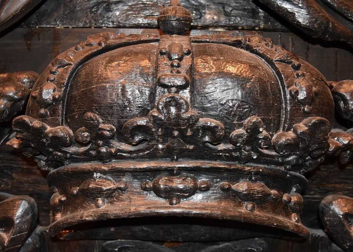 Skulpterad, kunglig krona. Undersidan är perspektiviskt framställd genom att kronans baksida gjorts högre än framsidan. Kronringen är upptill prydd med omväxlande större och mindre liljeliknande ornament. På själva ringen och på byglarna sitter fasettslipade "ädelstenar". Kronans baksida är slät.
Kronan är välbevarad.

Text in English: Royal crown. The underside is carved in perspective, achieved by carving the back of the crown higher than the front. The circlet is decorated at the top with lily-like ornaments of varying size. On the circlet itself and on the verticals are faceted "precious stones". The reverse side of the crown is smooth.
The sculpture is well preserved.