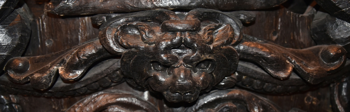 Broskornament. Ornamentet är utformat som ett kattdjursliknande huvud från vilket utgår "armar", dekorerade med blad och broskvoluter. Ändpartierna är förtjockade med elipsformade broskvoluter, blad och nedhängande flikar. Baksidan är slät.
Ornamentet är brutet på baksidans mittparti.

Text in English: Earshell ornament consisting of a cat-like head, from which extend "arms" decorated with ellipse-shaped pearlwork scroll leaves and downward-hanging leaf-blades with thickened ends. The reverse side is smooth.
The ornament is broken in the middle on the reverse side.