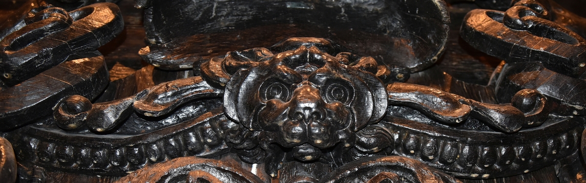 Broskornament. Ornamentet är utformat som ett kattdjursliknande huvud från vilket utgår "armar", dekorerade med blad och broskvoluter. Ändpartierna är förtjockade med elipsformade broskvoluter, blad och nedhängande flikar. Baksidan är slät.
Ornamentet är brutet på baksidans mittparti.

Text in English: Earshell ornament consisting of a cat-like head, from which extend "arms" decorated with ellipse-shaped pearlwork scroll leaves and downward-hanging leaf-blades with thickened ends. The reverse side is smooth.
The ornament is broken in the middle on the reverse side.