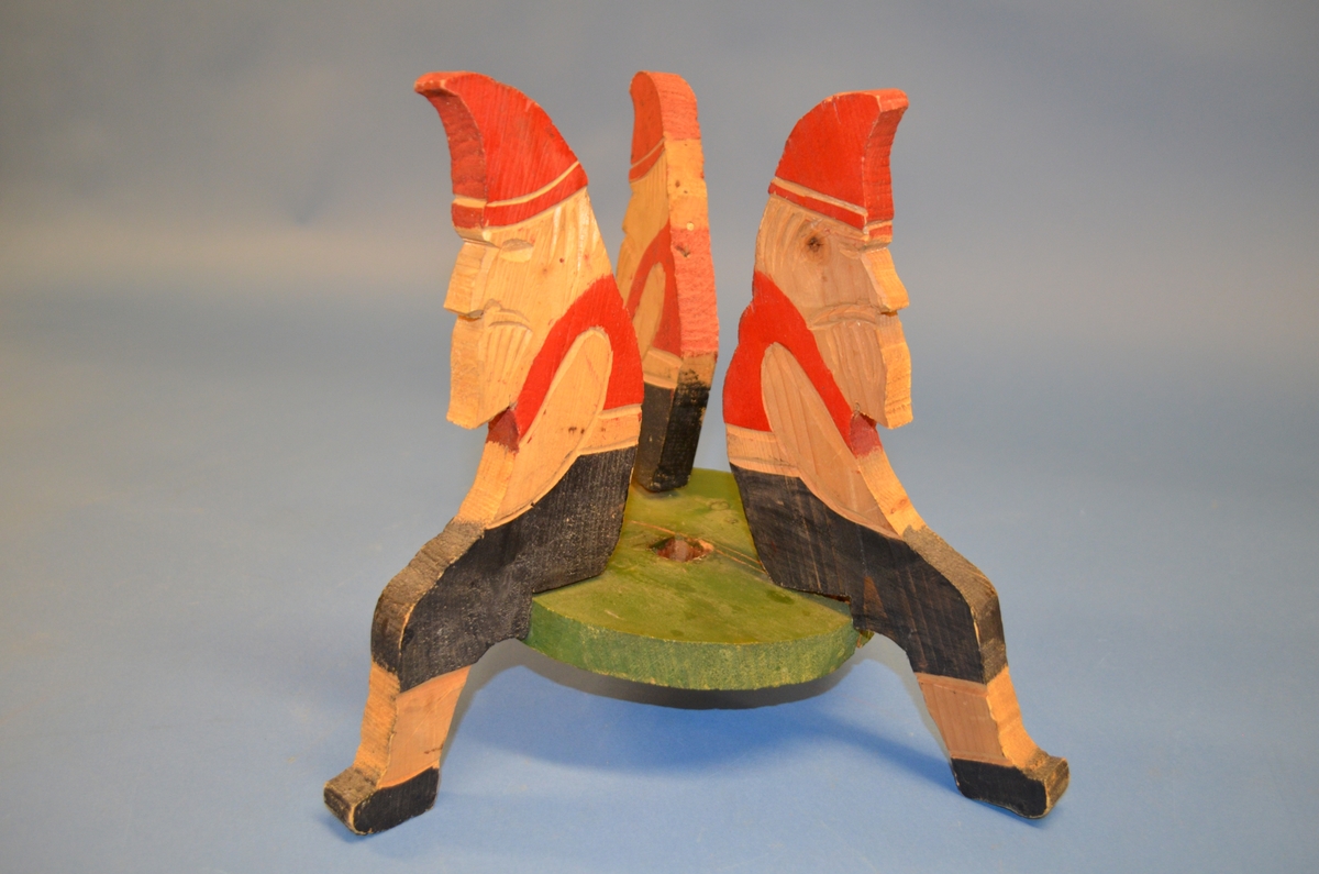 Hjemmelaget juletrefot av tre. Foten er satt sammen av tre støtter av hele trestykker formet som sittende nisser. I midten er et sirkelformet trestykke med hull for selve juletreet. Støtter og midtstykket er satt sammen med spiker. Juletrefoten er håndmalt. Nissene er malt i rødt og svart. Midtstykket grønt, med rosa underside.