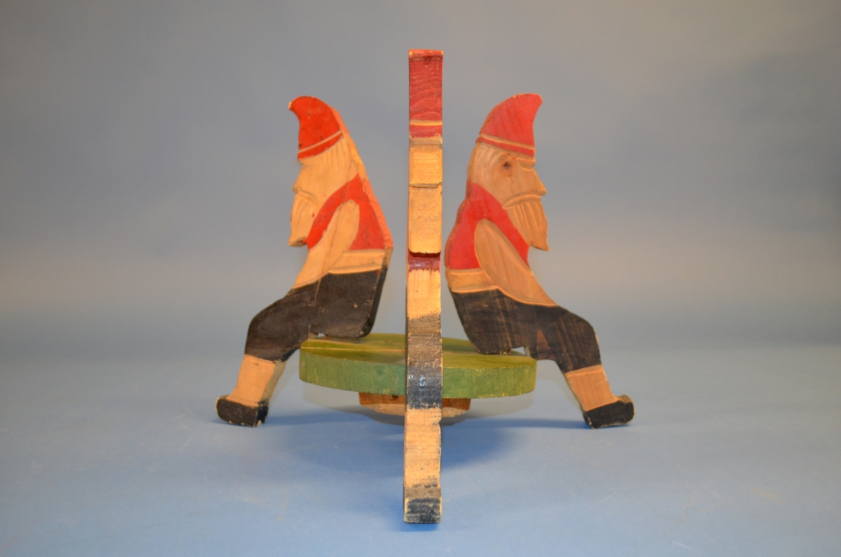 Hjemmelaget juletrefot av tre. Foten er satt sammen av tre støtter av hele trestykker formet som sittende nisser. I midten er et sirkelformet trestykke med hull for selve juletreet. Støtter og midtstykket er satt sammen med spiker. Juletrefoten er håndmalt. Nissene er malt i rødt og svart. Midtstykket grønt, med rosa underside.