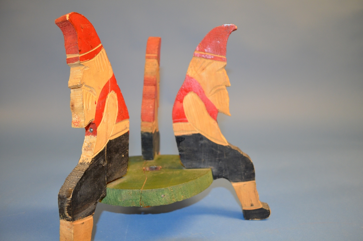 Hjemmelaget juletrefot av tre. Foten er satt sammen av tre støtter av hele trestykker formet som sittende nisser. I midten er et sirkelformet trestykke med hull for selve juletreet. Støtter og midtstykket er satt sammen med spiker. Juletrefoten er håndmalt. Nissene er malt i rødt og svart. Midtstykket grønt, med rosa underside.