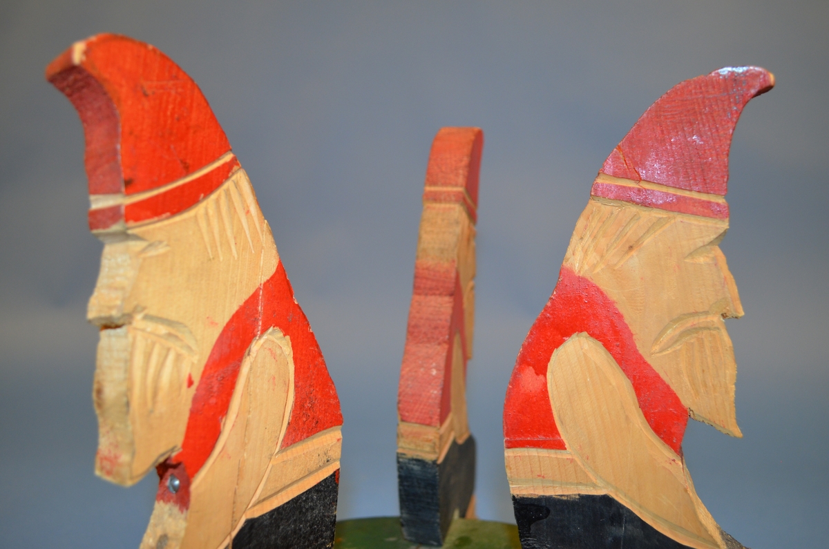 Hjemmelaget juletrefot av tre. Foten er satt sammen av tre støtter av hele trestykker formet som sittende nisser. I midten er et sirkelformet trestykke med hull for selve juletreet. Støtter og midtstykket er satt sammen med spiker. Juletrefoten er håndmalt. Nissene er malt i rødt og svart. Midtstykket grønt, med rosa underside.