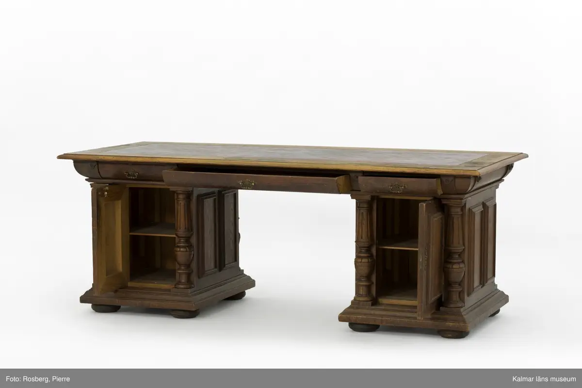 KLM 31941:1-3. Skrivbord. Tre delar. Stor fyrsidig skiva (:1) vilande på två hurtsar (:2-3), en vid var sida. Skivans ovansida är klädd i röd-svart skinn, omgivet av en ram i ekfanér. Skivan har i sargen sex utdragbara lådor, tre på varje långsida. Båda hurtsarna har två hyllplan och en dörr på var långsida. Hurtsarna är utformade med utskurna pilastrar som omger dörrarna samt på sidorna speglar. Hurtsarna står på platta kulfötter. Pulpet KLM 31942 har legat på skrivbordet.