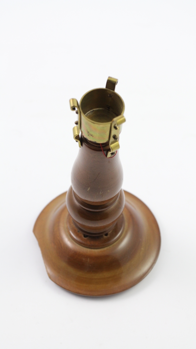 Bordlampe av tre. Tre små føtter i bunnen, bred base, metallstenger mellom basen og neste smale høye tre-element, metalldel til stearinlys eller lignende på toppen. En liten merkelapp henger i en rød tråd fra toppen, denne har muligens fungert som en prislapp.