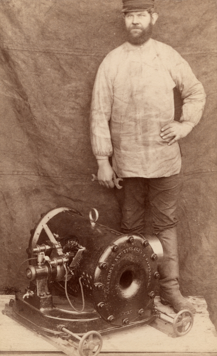 Visitkort för Elektriska Aktiebolaget föreställande verkstadsarbetaren Lars Johan Larsson vid elektrisk motor en dynamo. Fotograferat på vinden till Arboga Mekaniska Verkstad. Märkt på baksidan "Typ R. K-1300".