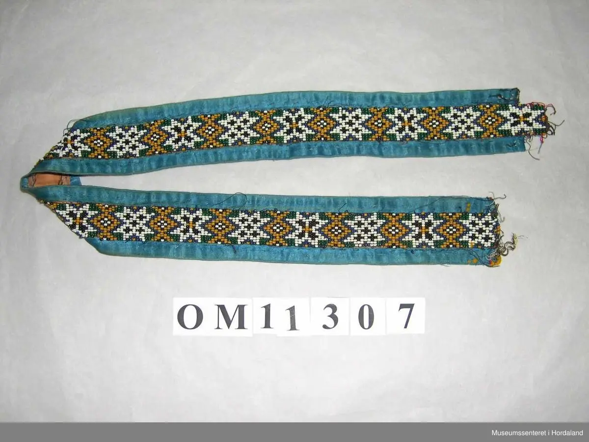 Perlebelte vove på perlegogn og montert på eit brunrosa bakstykke med blåe silkeband rundt kanten. Perledelen går litt utom kantbandet i eine enden. Motivet på perleveven er åttebladroser.