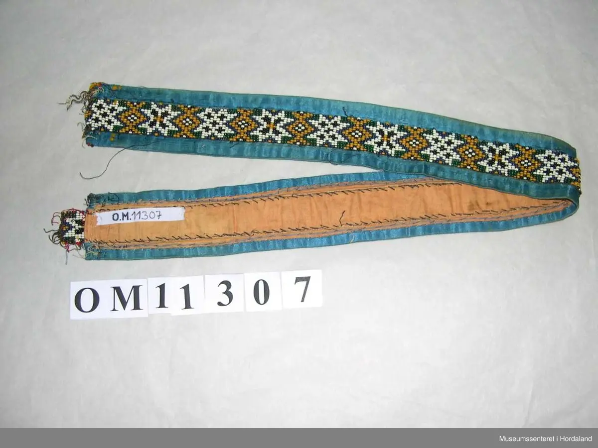 Perlebelte vove på perlegogn og montert på eit brunrosa bakstykke med blåe silkeband rundt kanten. Perledelen går litt utom kantbandet i eine enden. Motivet på perleveven er åttebladroser.