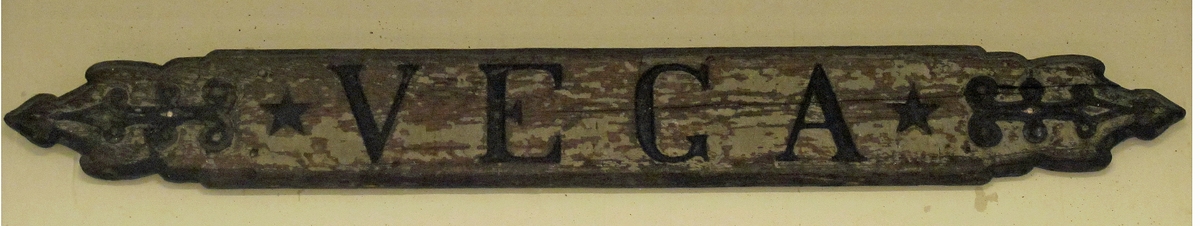 Namnbräda, "VEGA", inskrivet (rosartversaler) + femuddiga stjärnor, pilliknande dekor. Fasad kant, skulpterad i ändstyckena. Starkt avflagnad, grå färg. Text och dekor i svart.