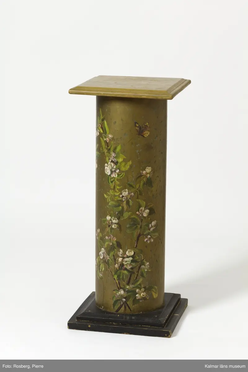 KLM 28494:113. Piedestal. Av trä och metall, plåt. Formad som en kolonn med skaft i bemålad plåt med guldfärg, samt med motiv av blommande fruktträdsgrenar i rosa och grönt, med en fjäril. Basplatta och överdel i kvadratisk form av trä, i svart respektive beige färg.