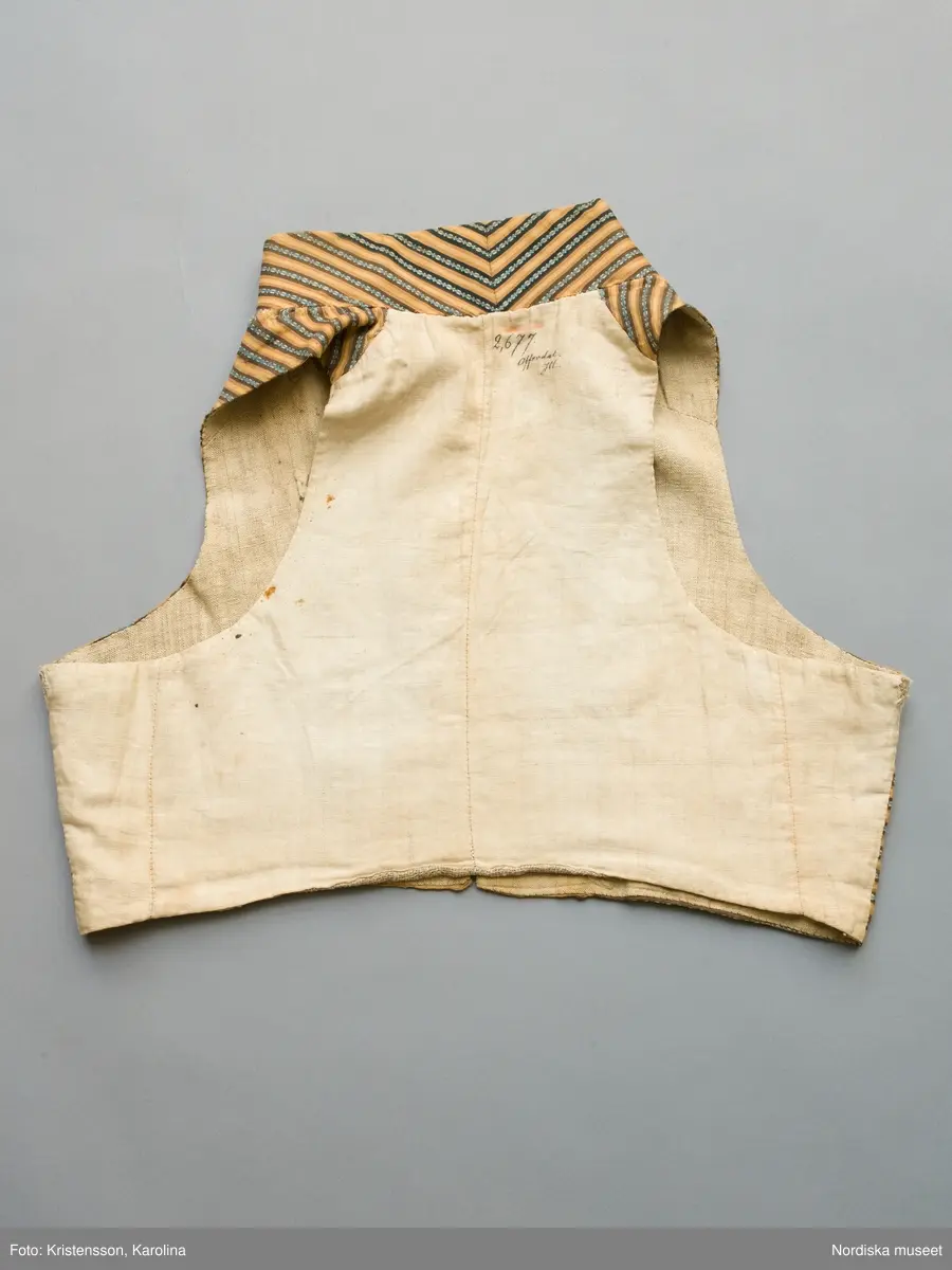 Väst i kort modell av randig bomullsrips i brunt och beige med invävd mönsterrand i den mörkbruna randen i ljusblått med vita "hoppor". Västen är tvärrandig med 2 framstycken och hög styvad  krage av samma tyg, ryggstycke av fin oblekt linnelärft, foder av grov och gles linnelärft i ryggen och finare i framstyckena, 2 stolpfickor, dubbelknäppning med knapphål på båda framstyckena och 6 par mässingsknappar med Ångermanlands landskapsvapen med 3 liggande laxar.
Tygsorten finns under benämningen: Västtyg i flera av 1820-30 talets tygprovsböcker och har enligt uppgift använts som brudgumsväst 1824
/Berit Eldvik  2005-11