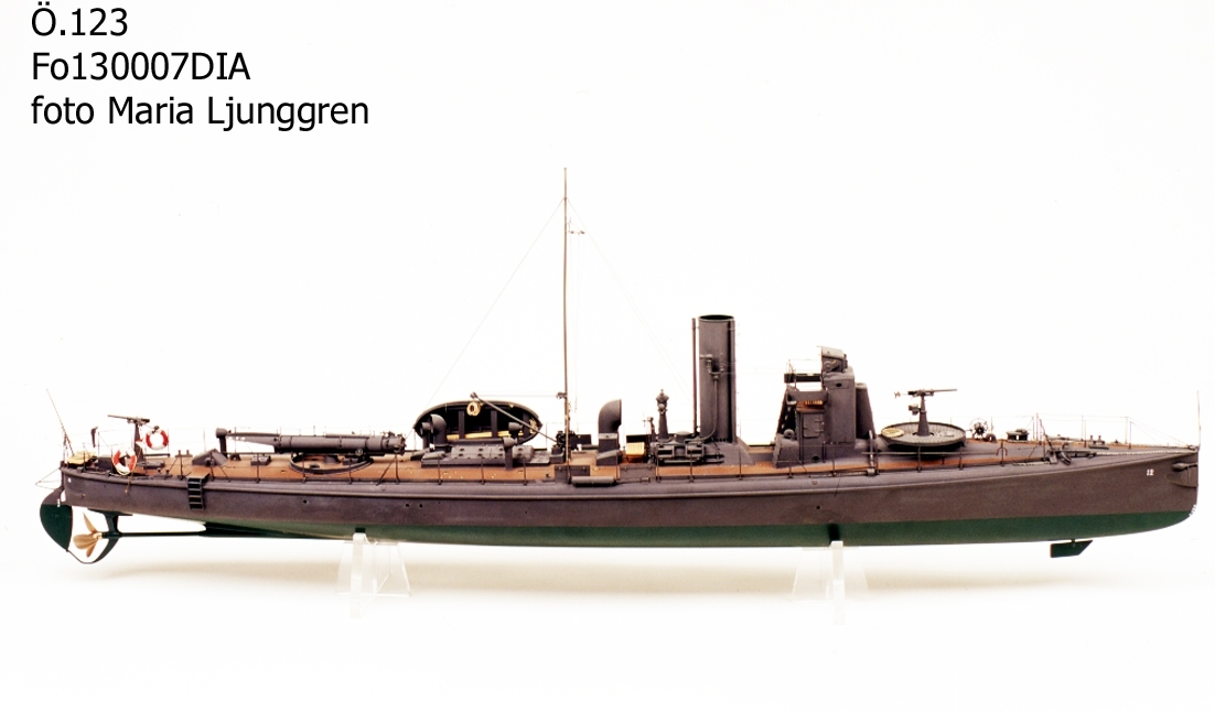 Fartygsmodell. Lamellbyggd, fullständig däcksinredning, för- och akterroder, metallpropeller. Riggad mast med vimpel, flaggspel med flagga, gösstake med gös. Gråmålad, grön botten, korkfärgat däck. 1 kanon. I stävarna märkt  '12” med vita siffror. 1 jolle med inredning, roder och 2 st åror. Stativ: 2 mässingspelare.