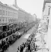 Förstamajdemonstration i Örebro den 1 maj 1937. Demonstrationståget går i riktning söderut på Drottninggatan. Det mycket långa förstamajtåget består av människor av olika slag. Några bär på röda fanor, plakat och banderoller. Åskådare kantar Drottninggatans båda trottoarer för att titta på demonstrationståget. Nikolaikyrkans torn kan skymtas ovanför hustaken till vänster i bilden. Närmast till höger står en kvinna på en balkong och tittar på evenemanget.