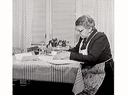 Maria och Knut Öberg sitter vid köksbordet. Maria skriver brev till sin bror i USA medan Knut läser tidningen. Bild 2: Porträtt av Maria när hon skriver brev.
