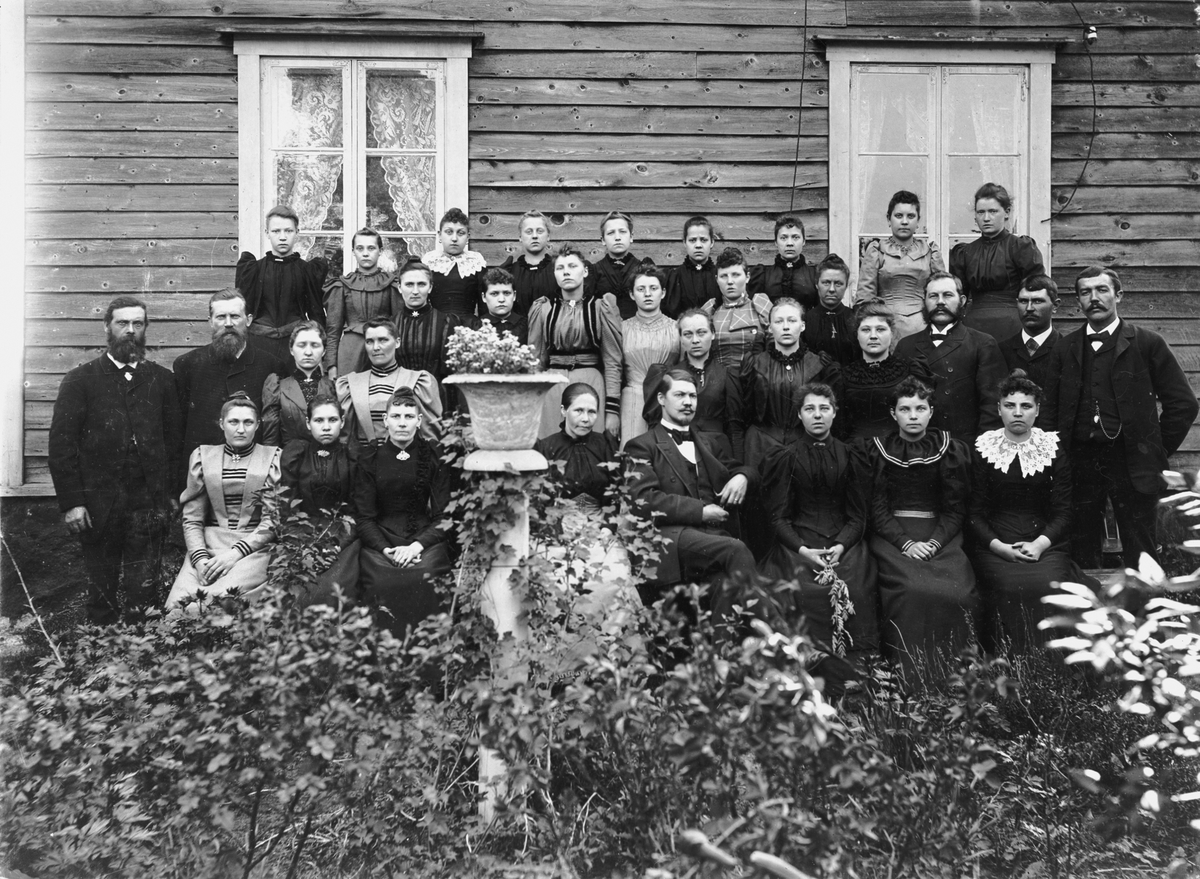 Större delen av personalen vid Liljeholmens Stubinfabrik samlad på pingstdagen 1894 framför fabrikörsstugans långvägg. Det ökande antalet "fabriksflickor" markerar företagets tillväxt. Ökningen av "fabriksflickor" tillkommer främst elektriska avdelningen. Mitt i första raden sitter verkmästare August Jansson.
