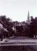 Kv. Klockaren 1, Storgatan 13, Kilanders. Kilanderska huset under byggnad. Färdigbyggt år 1900. Då inflyttade även postkontoret där från det lilla huset till vänster (se brevlådan på väggen) med ingång från gårdssidan. Postmästare var postexpeditör fru Maria Hammarstrand. I hörnet av Storgatan mellan Kilanders och lilla huset t. vänster låg poliskonstapel Högbergs låga envåningshus (ett stycke in på tomten), där sedan uppfördes hörnhuset inrymmande bokhandel, Skaraborgsbanken m.m. Till höger Jouvins hotell, och närmast kyrkan Westlins hus, där Kilander i många år hade sin manufakturaffär, tills hans nya hus blev färdigt.