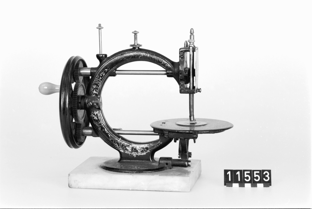 Handdriven symaskin på marmorplatta."The little Wanzer", för dubbeltrådig söm, med fram- och återgående skyttel enligt Howes patent. Märkt: R.M. Wanzer & Co. Patentees & Manufs Hamilton Canada West: Time utilizer. Trade (timglas) mark. Patented in America Gt Britain and the continent May 16 & June 17 1867".
Tillbehör: låda av trä med skjutlock och handtag, trälådans mått: 360x310x185,  Vikt:3,3kg, starkt sliten,  skyttel, 5 st spolar (2 saknas), 4 st fållvikare (saknas), 2 st ramar (?) för brodering(?), 2 st hållare (?) för brodering (?), 2 st kulor med filt (smörjkoppar ?).