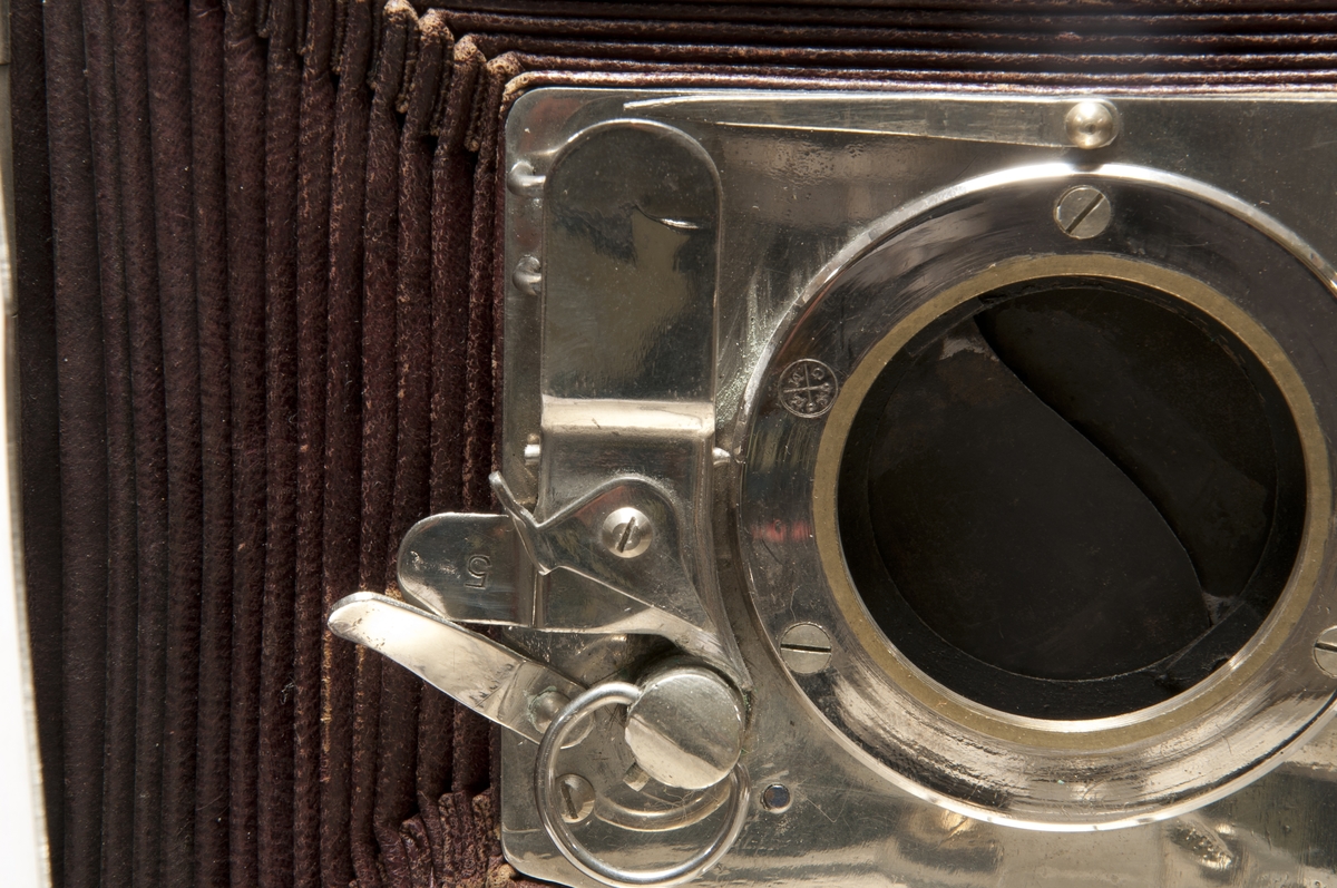 This was the first metal folding camera, the Model II. It was designed with a patent by H. Mader in 1888-1890. The camera was donated to the museum in January 1939 by Helmer Bäckström, who was a professor of photography. The camera is marked "5" in the slats of the bellows as well as with a round mark with the letters G M R and two sword-like characters. The lens is marked "Aplanal Invincible" and has "VI" engraved on the back at the threads. The camera comes with four cassettes, five plug-in apertures and a textile-clad case.