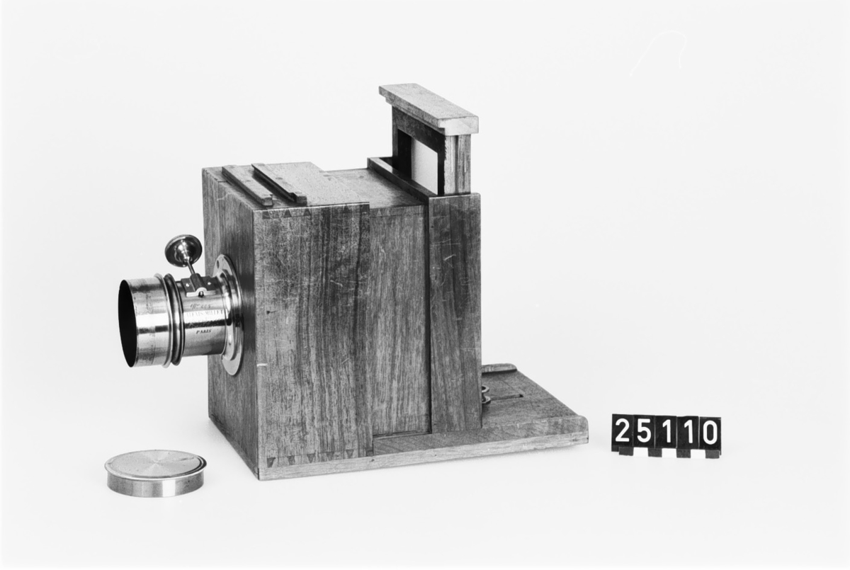Kamera, skjutlådemodell. Våtplåt 9,5 x 11,5. Objektiv: Alexis Millet, Opticien Bte'SGDG 160r.st:Martin Paris. Nr 605. Utrustning för fotografering på resor, jämte koffert. Lösa delar: kamera av trä med objektiv från Alexis Millet, Paris, nr. 605, objektivlock av mässing, visirskiva, 4 kassetter för våtplåtar 9 x 12 cm, bakstycke med visirskiva för stereobilder 8 x 8 cm, 2 kassetter för stereovåtplåtar, kärl av zinkplåt med lock (gångjärnet avbrutet), trattformat kärl av zinkplåt, mindre kuvert av glas (spräckt och lagad, troligen av givaren), större kuvert av glas, torkställ av trä, 2 skivor av trä med spår för plåtar, kopieram för max 16 x 20 cm, vikbar skiva av trä (för beredning, troligen av våtplåtar), liten bordskiva med kantlist (passar till stativet TM 25.111,fodral av trä med 12 st. våtplåtnegativ 9 x 12 cm (porträtt och skulpturer), fodral av trä med 12 st. sämre negativ, fodral av trä med 10 st. blanka och  skrapade glasplåtar, diverse plåtar i paket (i ask ny 1950), fodral av trä med 11 st. stereovåtplåtar, varav 3 preparerade.
Tillbehör: Koffert.