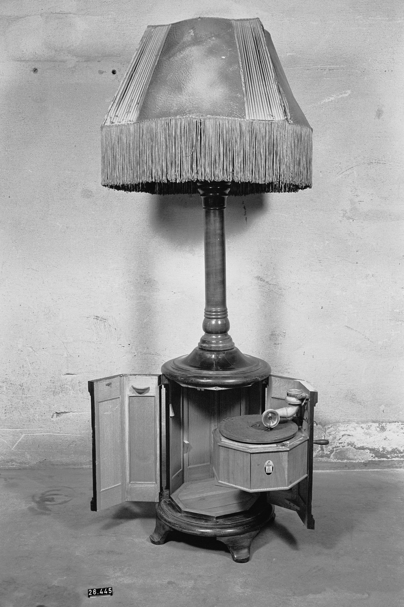Stor golvlampa med grammofon. Ljudet leds i piedestalen till ljudöppning under lampskärmen. Åttkantigt underskåp av mahogny och mahognybetsad bok, med inredning av ek, innehållande fjäderdrivet grammofonverk och ljudarm av trä. I ytterväggen finns hål för vev. Ihålig pelare, upptill avslutad med pyramidformad ljudtratt av trä med fyra glödlampor. Däröver en fyrkantig (buktig) lampskärm av mönsterpressat "pergamentpapper" och siden med silkesfrans. Skärmen (spec. sidenet) mycket skadad. Skärmens diameter: ca. 800 mm. Grammonfonen stannar själv då skivan är slutspelad. Nu befintlig elektrisk ljuddosa monterad senare i stället för ursprunglig membrandosa (som saknas).
Tillbehör: Vev.