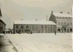 Stora torget. Omkring 1912. Den s.k. Thulins-tomten vid Stora torget. Utanför sin butiksdörr står gamle glasmästare  A. Larsson, därnäst (åt höger) är Kristinedals fabriksbod, och ännu längre till höger synes Hilma Brelins vita skylt. Ingången till hennes affär var från gårdssidan. I gatuhörnet står fastighetens ägare byggmästare Emil Gustafson (fr.Motala).