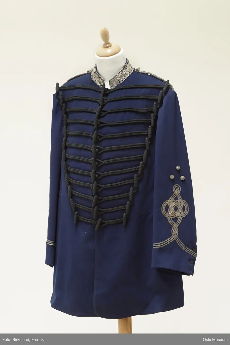 a) Våpenkjole av blått klede m. trangt liv. Kragen brodert med sølvstresser, flettede epauletter. Snorbesetning av sort silke, 3 knapperader. Fòret med sort satin. H: 97
b) Benklær av blått klede med sølvgaloner. L: 117
C) Kepi m. 3 rader sølvstresser om pullen, kokarderosett .

1875. Har tilhørt rittermester Hans Fredrik Grüner. Gave fra fru Charlotte Grüner 1913.