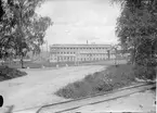 Hagfors bruk. Elektriska hyttan. Husen av rött tegel. Stora huset med tegeltak. Små husen med svart papptak, dörrarna mörkröda, rött staket. Fabriksskorstenarna av rött tegel.