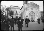 Skioptikonbild från institutionen för fotografi vid Kungliga Tekniska Högskolan. Bilden är troligen tagen av John Hertzberg under en resa i Europa. Hertzberg blev docent i fotografi 1921vid Kungliga Tekniska Högskolan i Stockholm. Folkliv vid kyrkan.