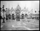 Skioptikonbild från institutionen för fotografi vid Kungliga Tekniska Högskolan. Motiv från Markusplasten (Piazza di San Marco) i Venedig. I bakgrunden, centralt i bilden, syns Markuskyrkan (Basilica di San Marco). Bilden är troligen tagen av John Hertzberg under en resa i Europa.