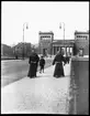 Skioptikonbild från institutionen för fotografi vid Kungliga Tekniska Högskolan. Motiv föreställande munkar promenerande framför Propylaea i München. Proypylaea är en stadsport vid västra sidan av Köningsplatz. Den stod färdig 1862. Bilden är troligen tagen av John Hertzberg under en resa i Europa.