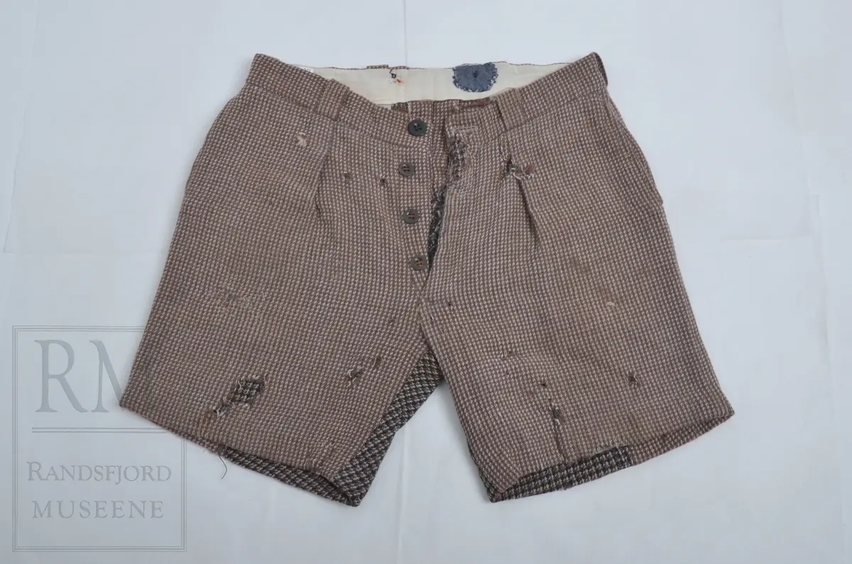 Kypertvevet ullstoff, tweed. Linningen foret med kypertvevet hvitt bomullslerret. Linningen har 6 stolper til belte. To patentknapper bak. Gylf med tre knapper. Stikklomme i sidesømmene. Lomme på høyre side bak. Stor lapp i baken. Hull og stopping- Legg og press
