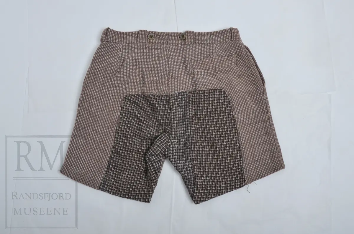 Kypertvevet ullstoff, tweed. Linningen foret med kypertvevet hvitt bomullslerret. Linningen har 6 stolper til belte. To patentknapper bak. Gylf med tre knapper. Stikklomme i sidesømmene. Lomme på høyre side bak. Stor lapp i baken. Hull og stopping- Legg og press