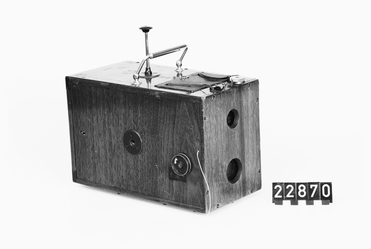 Lådkamera av trä, för plåtar 6.5 x 10 cm, i fodral av läder, med invändig plåtväxling. S.k. fallplåtskamera. Originell plåtväxling (troligen patent). Stor brilljantsökare med uppfällbart bländskydd, roterande skivbländare, skilda knappar för tids- och momentexponering samt plåträknare. Ögonblicksslutare. 30 st metallplåtar
Tillbehör: Kamera, fodral av läder samt 30 plåthållare av svart bleckplåt.