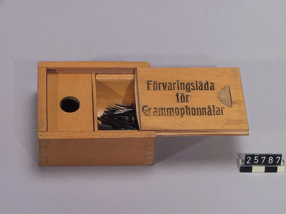 Ask av trä till förvaring av grammofonnålar. Med skjutlock med text: "Ask för Grammophonnålar. Ett fack med halvcylindrisk botten för obegagnade nålar, ett fack med lock med hål för begagnade".