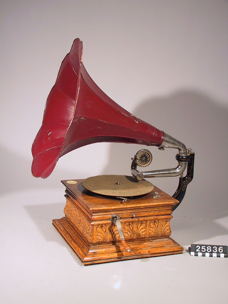 Grammofon med tratt av rödlackerad plåt (tratt TM 34.449). Ljuddosa "Exhibition", tillverkad i USA av Gramophone & Typewriter Ltd (London Berlin Paris). Lådan och troligen verket av Deutsche Gramophon AG. Lådan stämplad i botten "DGAG 7484 Pev K". Skylt med en "skrivande ängel" på lådan. Enkelfjädrigt verk.