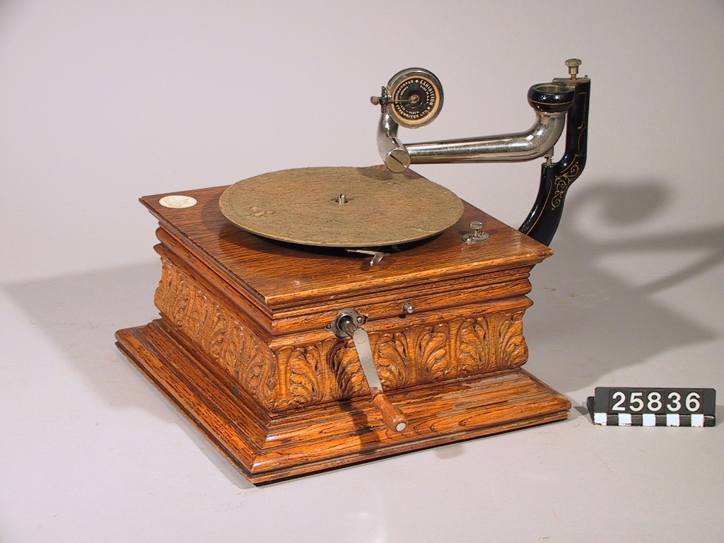 Grammofon med tratt av rödlackerad plåt (tratt TM 34.449). Ljuddosa "Exhibition", tillverkad i USA av Gramophone & Typewriter Ltd (London Berlin Paris). Lådan och troligen verket av Deutsche Gramophon AG. Lådan stämplad i botten "DGAG 7484 Pev K". Skylt med en "skrivande ängel" på lådan. Enkelfjädrigt verk.