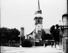 Skioptikonbild med motiv av Stadtpfarrkirche Sankt Kilian, Fladungen, vid sidan ett torn i stadsmuren.
Bilden har förvarats i kartong märkt: Vårresan 1910. Fladungen 7. X.