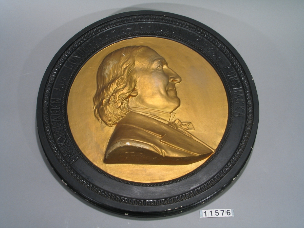 Medaljong i gips över August Lundeberg. Höger profil, guldbronserad, runtom text: "Bruksegaren Aug. Lundeberg x 18 31/10 10 å Ängsö + 18 25/9 85 å Forsbacka". Signerad: "C.J. Dyfverman år 1887"
Tjocklek 85 mm.