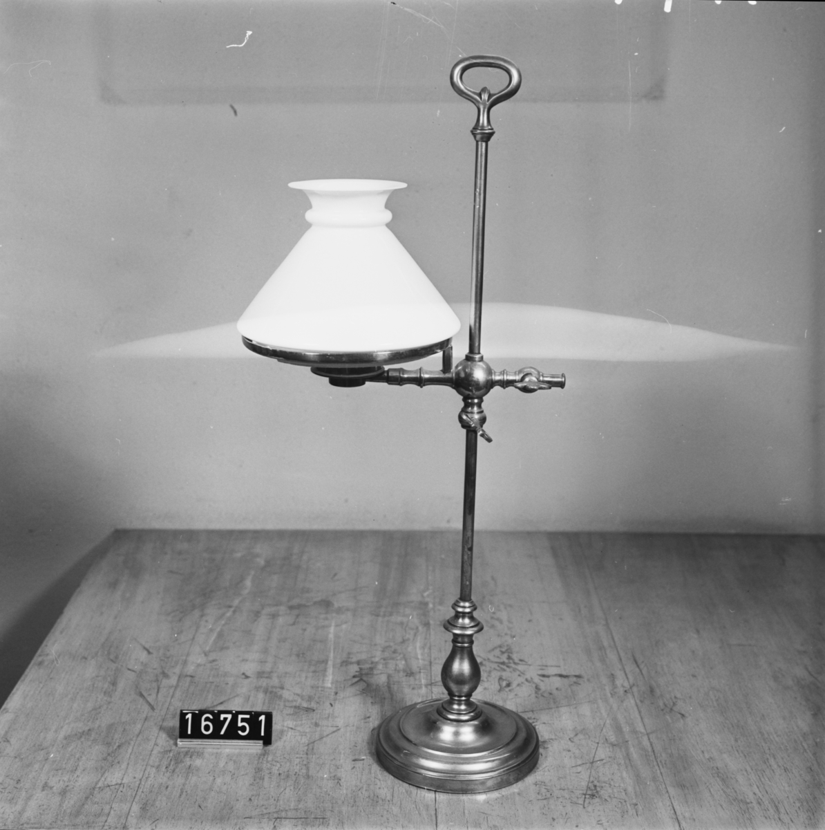 Bordslampa med höj- och sänkbar brännare av porslin samt lampkupa och kupring. Lampkupa: TM 18.136. En flisa borta ur kupans nedre hals. Lampglas saknas dock.
Tillbehör: Lösa delar: lampfot, kupring, lampkupa.