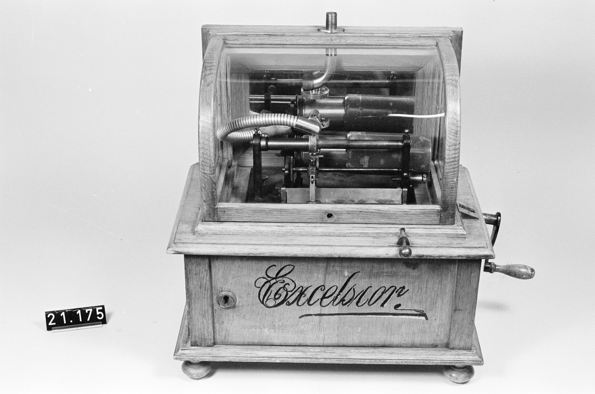 Fonograf, "Excelsior", med myntautomat, för normal 2-minuters vaxrullar. Vaxrulle: längd: 500 mm, bredd: 250 mm, höjd: 420 mm. Under huv av glas i ram av trä. Verkets fjäder är brusten och tratt saknas, har varit snett uppåt mot den lyssnande, fästad genom böjt skarvstycke. En sliten vaxrulle medföljer.