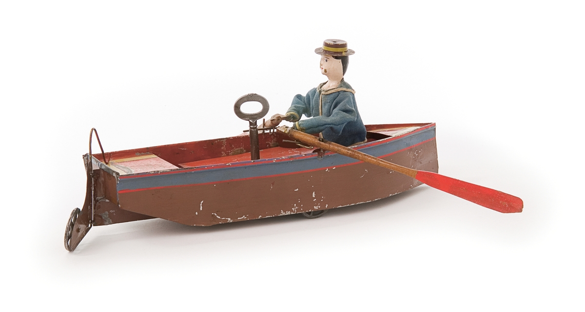 Mekanisk leksak av plåt, går på dolda hjul. Brunlackerad (handmålad) med blå reling och rött däck över ett fjäderverk som påverkar en matrosfigur i blå tygjacka, manövrerar ett par åror.
Tillbehör: Nyckel.